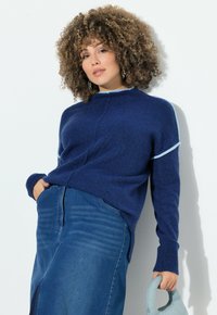 Frau mit lockigem Haar, die einen blauen Pullover und einen Jeansrock trägt, eine hellblaue Handtasche hält und vor einer weißen Wand steht.