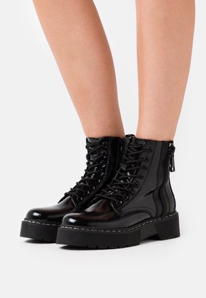 Botas de tobillo brillantes de cuero negro con cordones y suelas gruesas, llevadas sobre piernas desnudas, mostradas contra un fondo blanco.