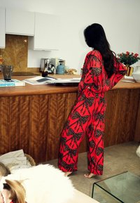 Punase mustriga kombinesoon mustade lehemotiividega, lõdva istuvusega, pika varrukaga, seisab puidust leti kõrval, millel on erinevad köögitarbed.