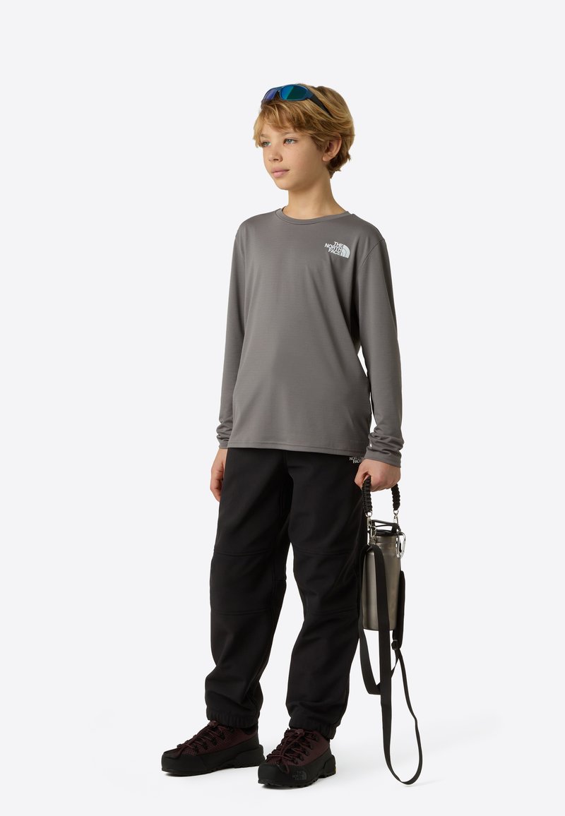 Sivá košeľa s dlhým rukávom s logom, čierne nohavice a tmavé topánky. Doplnky zahŕňajú držiak na fľašu a slnečné okuliare s modrým odtieňom.