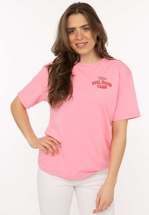 Junge Frau mit langen braunen Haaren, die ein pinkes T-Shirt mit dem Schriftzug "FEEL GOOD CLUB" und weiße Hosen trägt, lächelnd mit der Hand in der Hüfte.