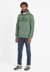 Green cotton hoodie with a large gray "TOG24" logo, featuring a kangaroo pocket and gray drawstrings, paired with dark blue jeans and mixed-color sneakers.