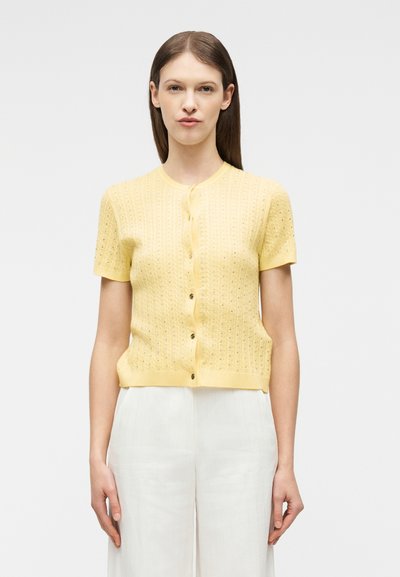 Γυναίκα με ίσια καστανά μαλλιά που φοράει κοντομάνικο πλεκτό ζακετάκι με κουμπιά σε κίτρινο χρώμα και άσπρο παντελόνι, που στέκεται μπροστά σε απλό φόντο.