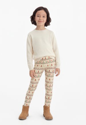 Barn med kort mørkt hår iført en kremfarget genser, mønstrede leggings med bamser og trær og beige støvler på hvit bakgrunn.