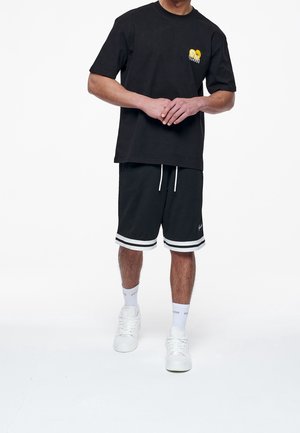 Camiseta negra de algodón con gráfico amarillo en el pecho, combinada con pantalones cortos negros con rayas blancas y cordón. Zapatillas blancas completan el conjunto.