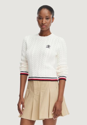 Donna che indossa un maglione bianco a trecce con polsini e orlo a righe rosse, bianche e nere abbinato a una gonna plissettata beige.