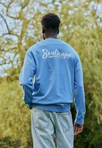 Homme portant un sweat-shirt bleu "Soulscape" et un jean clair, debout en plein air avec des arbres en arrière-plan, vu de dos.