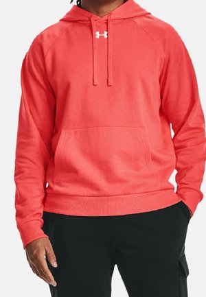 Mężczyzna w jasnokoralowej bluzie z kapturem z przednią kieszenią kangurka, białym logo Under Armour oraz czarnych spodniach z boczną kieszenią.