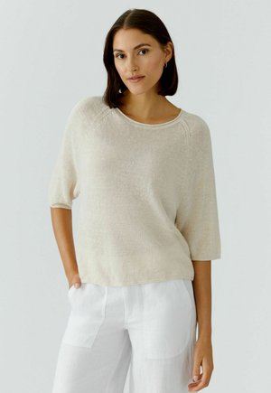 Vrouw met donkerbruin haar, gekleed in een crèmekleurige gebreide top met ellebooglange mouwen en een witte broek, staand met één hand in haar broekzak.