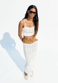 White crochet crop top and long skirt with intricate patterns, complemented by a tan belt. Model wears dark sunglasses and neutral-toned footwear.