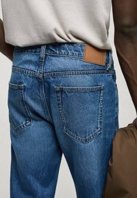 Blue denim jeans with a slightly tapered fit, featuring a brown leather patch on the waistband and two back pockets.
