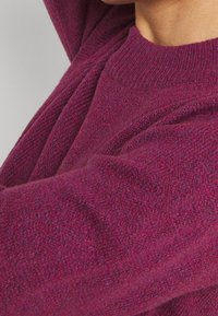 Fioletowy sweter z dzianiny o fakturowanej tkaninie. Posiada okrągły dekolt i długie rękawy. Materiał wygląda na miękki i ciepły.