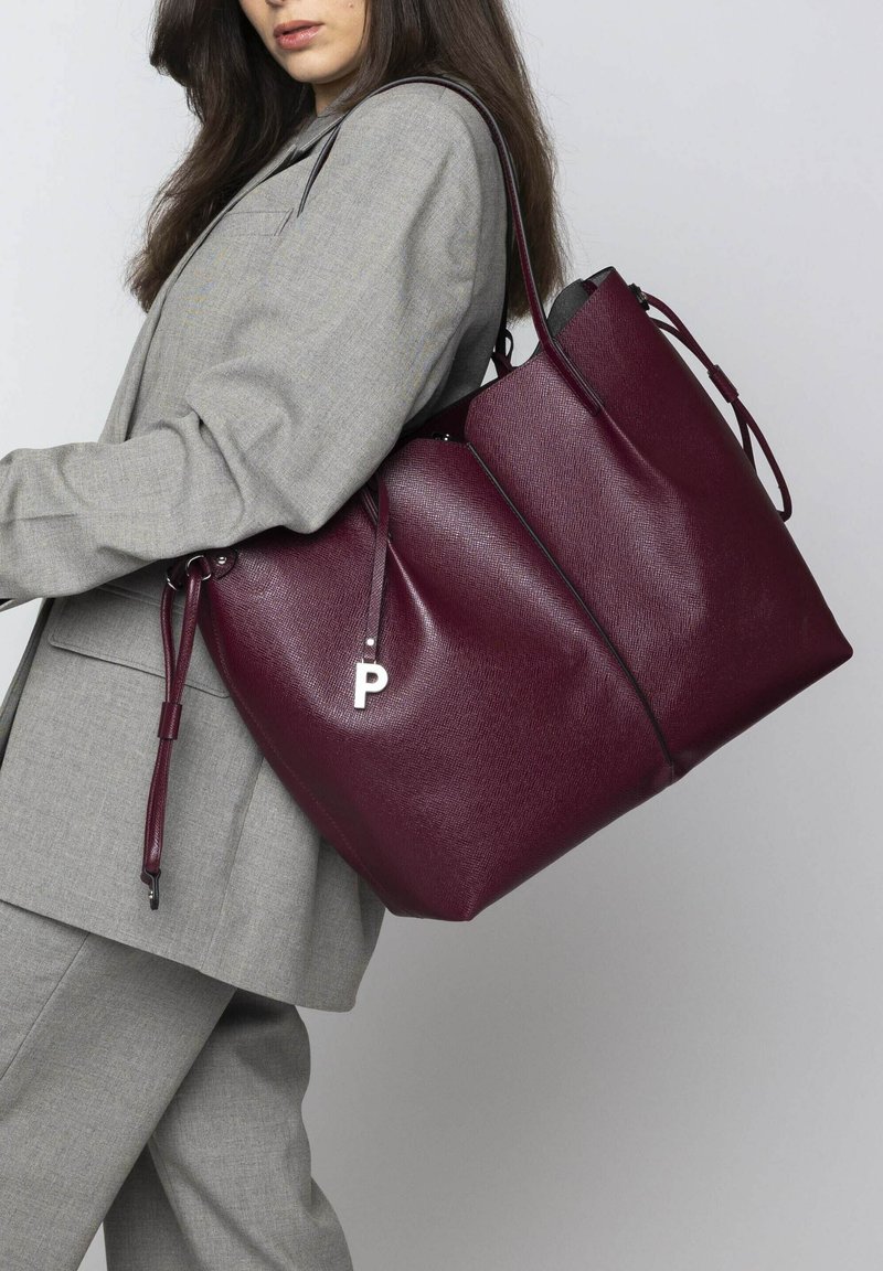 Sac tote en cuir bordeaux avec une finition texturée, orné d'un pendentif en forme de lettre "P" en argent et de sangles ajustables. Design large et spacieux.