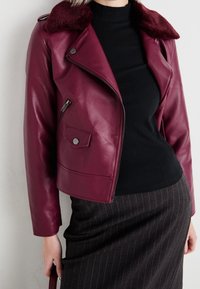 Burgundy läderjacka med fårskinnskrage, med fickor som stängs med dragkedja och tryckknappar, bärs över en svart ribbad tröja.