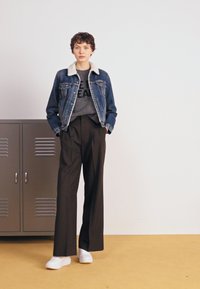 Jeansjacka med fårskinnskrage, grå grafisk t-shirt, högmidjade svarta byxor och vita sneakers, stående mot ett grått skåp.