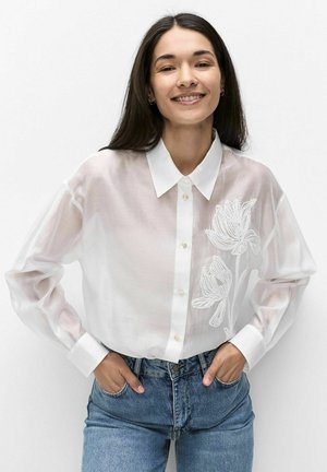 Vrouw die glimlacht met lang donker haar, draagt een doorschijnende witte blouse met bloemrijke borduurwerk en blauwe spijkerbroek, handen in haar zakken, staand tegen een witte achtergrond.