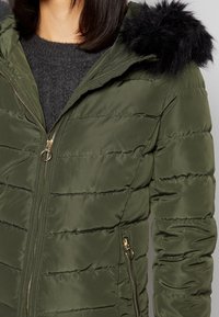 Femme portant une veste matelassée vert olive avec bordure de capuche en fausse fourrure noire et détails de fermeture éclair dorés sur un pull gris foncé.