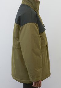 Olivgrüne und schwarze Jacke mit hohem Kragen, Fronttasche und boxy Silhouette. Hergestellt aus einem glatten, strapazierfähigen Stoff.