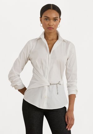 Vrouw in een wit overhemd met lange mouwen en een zilveren kettingceintuur, gecombineerd met zwarte broek, staande tegen een witte achtergrond.