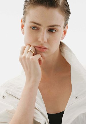 Anillo de oro con un diseño curvado que se lleva en la mano, combinado con una chaqueta blanca de cuello alto y una camiseta negra. Fondo limpio y minimalista.