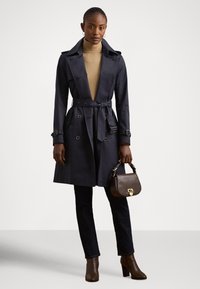 Ναυτικό παλτό στρατιωτικού τύπου με ζώνη στη μέση, διπλό κούμπωμα κουμπιών και πλαϊνές τσέπες, συνδυασμένο με σκούρο τζιν και καφέ μπότες με τακούνι.