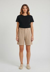 Mujer de pie con camisa negra de manga corta, pantalones cortos beige de talle alto y zapatillas beige, posando contra un fondo claro y sencillo.