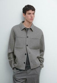 Grå uldblandet jakke med krave, to brystlommer og sorte knapper, kombineret med en mørk skjorte og lysegrå bukser.