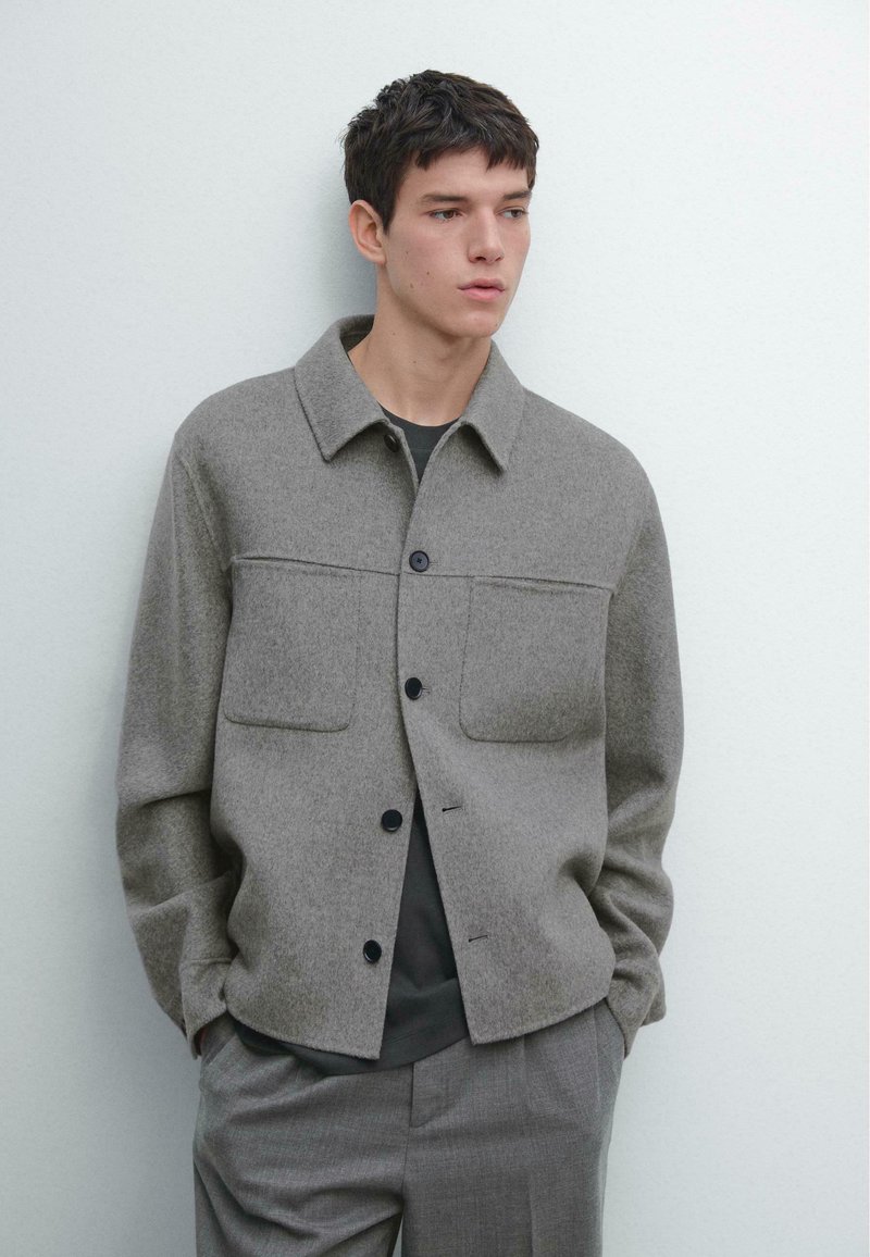 Grå uldblandet jakke med krave, to brystlommer og sorte knapper, kombineret med en mørk skjorte og lysegrå bukser.