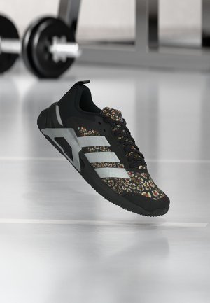 Pantof sport negru cu model floral, dungi gri și talpă groasă, plutind deasupra podelei reflectorizante din sala de sport, cu gantere neclare în fundal.