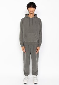 Grå bomulds hoodie med en lomme foran og snøre i hætten, parret med matchende grå sweatpants og hvide sneakers. Glat tekstur.