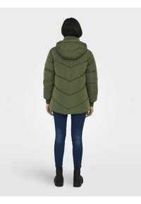 Storos pūkinės striukės tamsiai alyvinės žalios spalvos su gobtuvu, chevron formos siuvimu ir ribomis rankogaliais. Dėvima kartu su mėlynais džinsais ir juodomis aulinėmis batais.