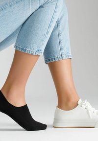 Białe płócienne trampki z zaokrąglonym noskiem i sznurowanym zapięciem obok czarnej, niskiej skarpetki. Widoczne jasnoniebieskie krótkie dżinsy. Gładki odcień skóry.
