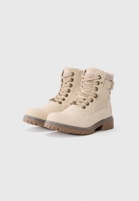 Botas de tobillo beige hechas de un material similar al ante, con ojales de metal, cordones y una correa. La suela es de goma texturizada para mayor tracción.