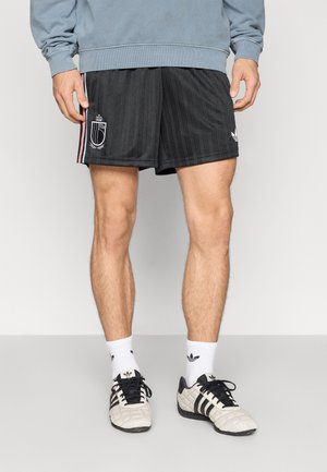 Μαύρα αθλητικά σορτς με κατακόρυφες ρίγες, λογότυπο και ροζ λεπτομέρειες. Φορεμένα με λευκές κάλτσες και μαύρα παπούτσια με υφασμένη σχεδίαση.