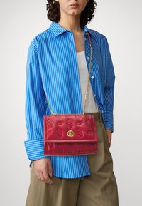 Borsa a mano rossa in rilievo con una chiusura dorata, dotata di una tracolla a catena. Abbinata a una camicia a righe blu e pantaloni beige. Dettagli della superficie testurizzati.