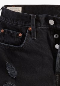 Gros plan sur un jean en denim noir avec poche avant, rivets en cuivre, coutures visibles, détail déchiré et braguette à boutons argentée partiellement ouverte.