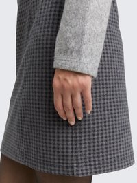 Mano appoggiata su una gonna a quadri grigia e blu scuro con una manica di lana grigia chiara visibile.