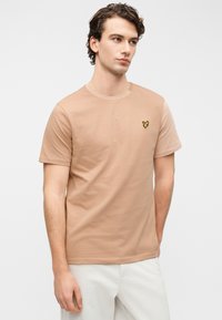 Ανοιχτό καφέ βαμβακερό t-shirt με κοντά μανίκια, που διαθέτει ένα μικρό, κεντητό κίτρινο λογότυπο πεταλούδας στην αριστερή πλευρά του στήθους.