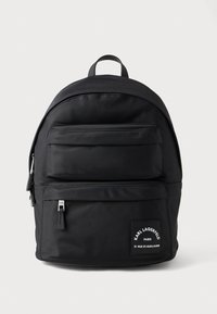 Musta nailonireppu pyöreällä muodolla, etutaskussa vetoketju, hillitty brändäys ja ylhäällä kantokahva. Sileä pinta.