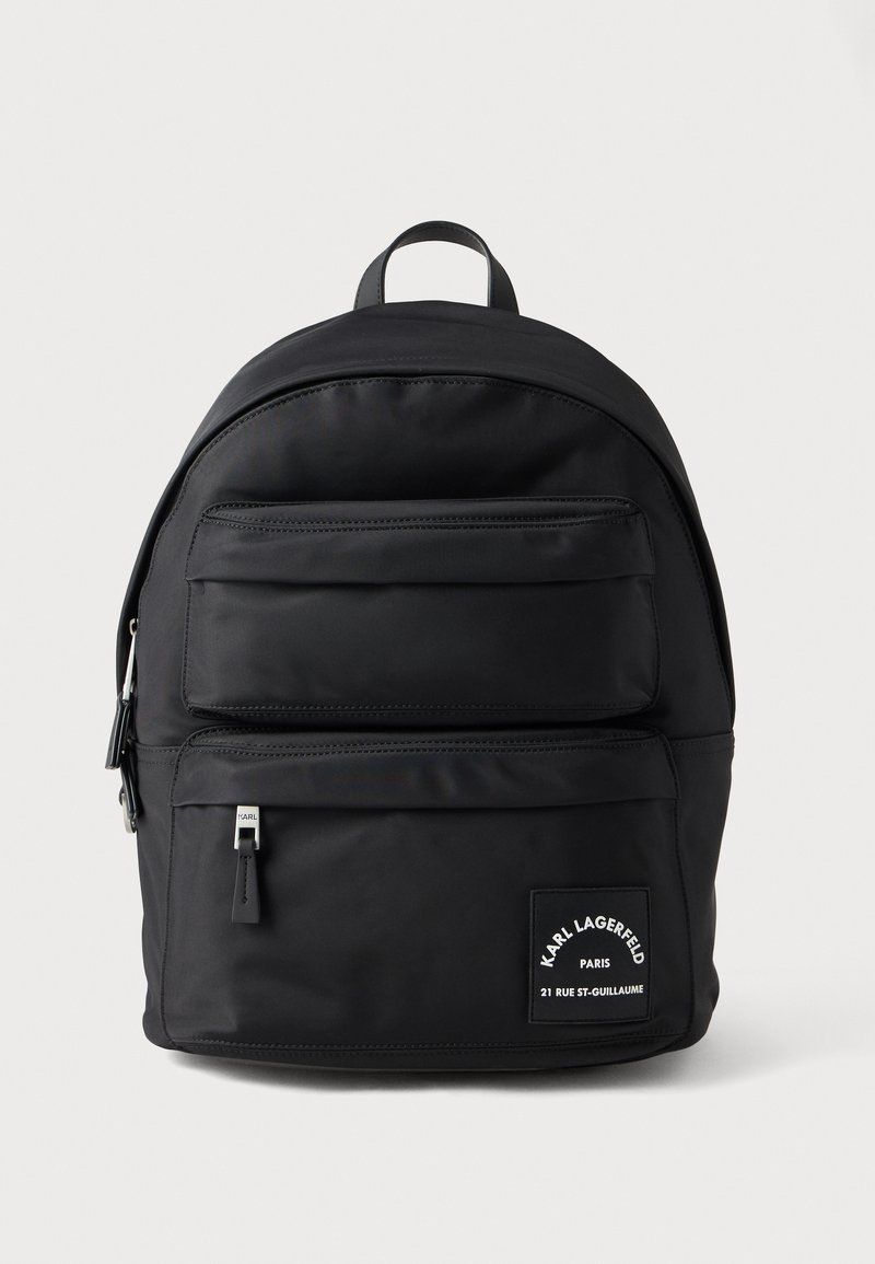 Musta nailonireppu pyöreällä muodolla, etutaskussa vetoketju, hillitty brändäys ja ylhäällä kantokahva. Sileä pinta.