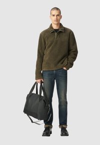 Pull en polaire vert olive avec un col zippé à mi-hauteur, associé à un jean bleu et des chaussures noires, tenant un sac fourre-tout noir à deux poignées.
