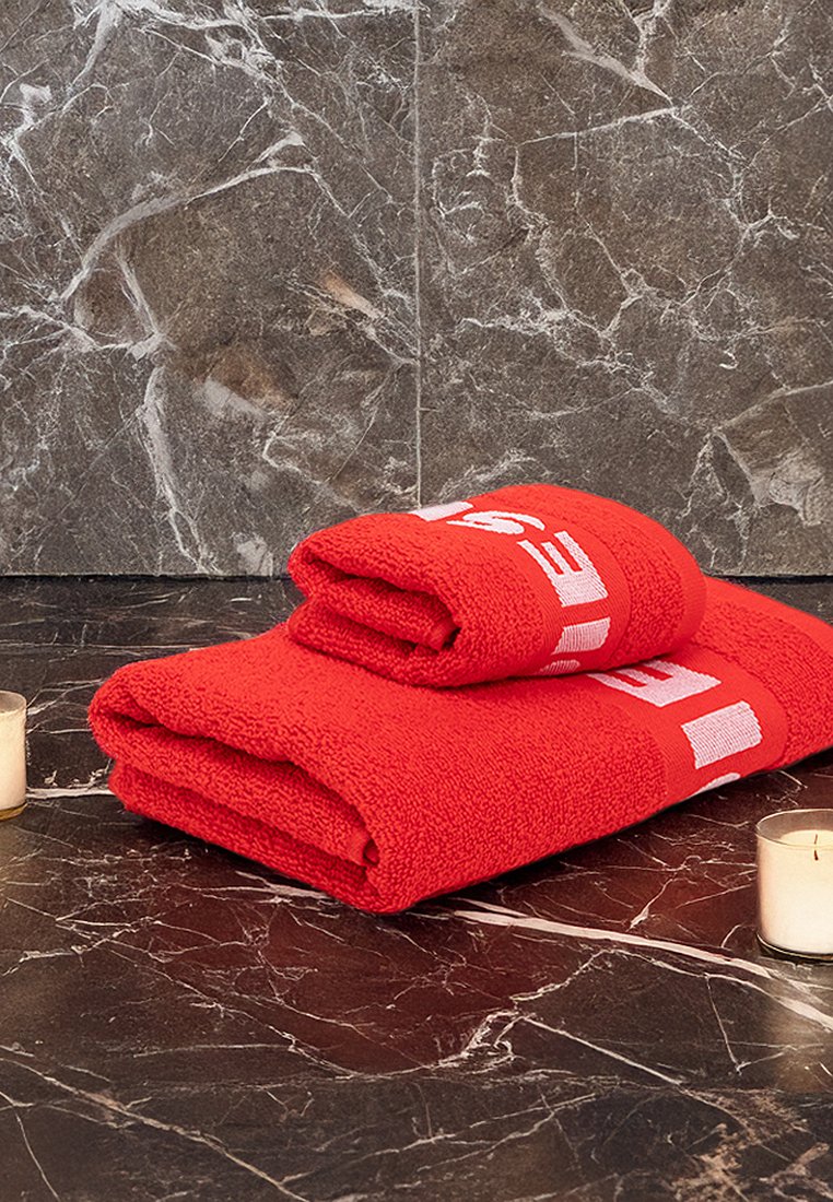 Toallas de algodón rojas apiladas sobre una superficie de mármol, con una textura de tela y letras blancas prominentes. Pequeña vela junto a las toallas.