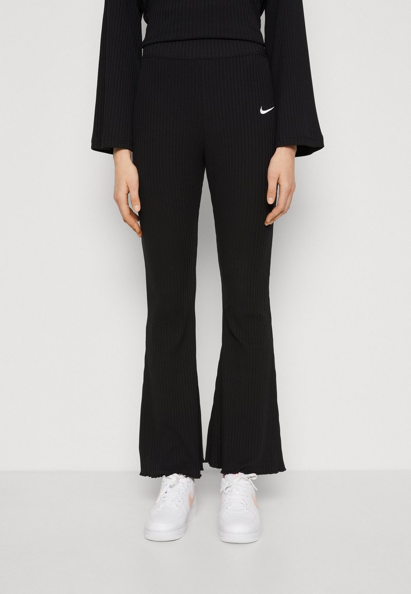 Kvinna i svarta ripplade flare byxor med vit Nike-logotyp och vita sneakers, som står mot en enkel ljus bakgrund.