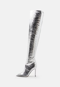 Bota plateada por encima de la rodilla hecha de material brillante con punta afilada y tacón alto de estiletto; presenta una textura suave con leves arrugas.