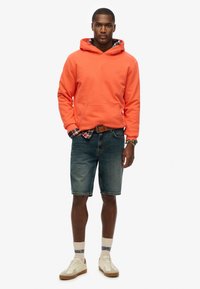 Sudadera naranja con capucha y un bolsillo tipo canguro, combinada con pantalones cortos de denim azul. Se pueden ver zapatillas de color claro y acentos de calcetines blancos.