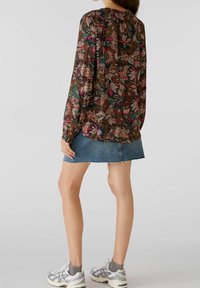 Femme portant un blouse à manches longues à fleurs, une jupe en denim, des chaussettes grises et des baskets blanches, se tenant contre un fond uni.