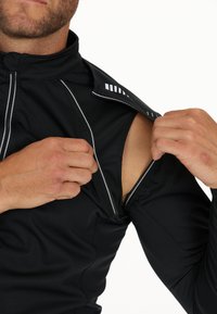 Svart atletisk topp med hög krage, med en dragkedja som öppnas och avslöjar bar hud. Reflekterande detaljer längs dragkedjan för bättre synlighet.