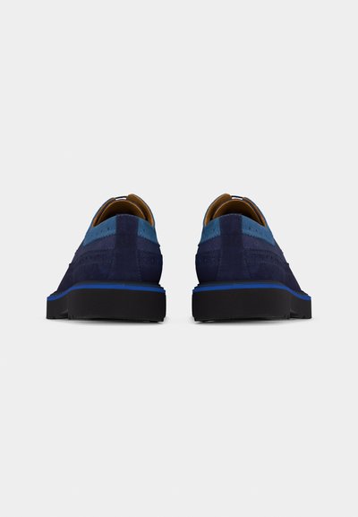 Paire de chaussures décontractées en daim bleu marine et bleu clair avec semelles noires, vues de dos sur un fond uni gris clair.