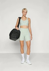 Fiatal nő rövid szőke hajjal, világos zöld sportmelltartóban és sortban, fehér sportcipőben és zokniban, a vállán fekete edzőtáskával.