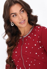 Chaqueta roja con cremallera y tejido texturizado, adornada con perlas falsas y un cuello con detalles de brillantes. Combinada con pendientes grandes de aro.